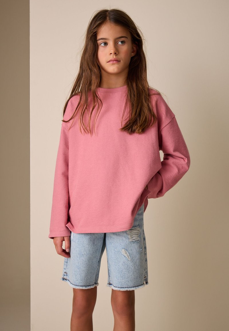 Jeune fille aux longs cheveux bruns portant un pull rose ample et un short en jean bleu clair déchiré, debout avec une main dans la poche.