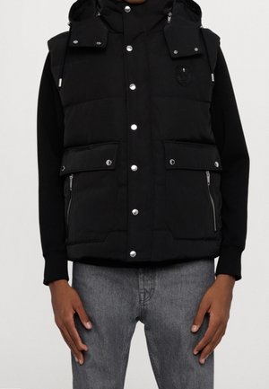 Mężczyzna ubrany w czarną pikowaną kamizelkę z kieszeniami na guziki i suwak, na czarnej koszuli z długim rękawem oraz wyblakłych szarych dżinsach, stojący na tle jednolitego tła.