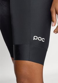 Sorte sykkelskotter med en glatt tekstur som har en hvit "poc"-logo på siden. Designet er tettsittende og midt-lår lengde.