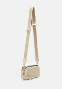 Petit sac bandoulière beige avec une large sangle réglable et une fermeture éclair dorée, présenté sur un fond clair uni.