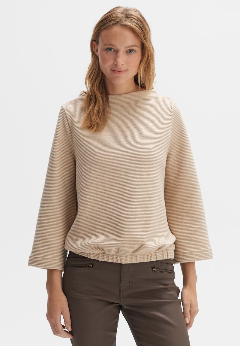 Beige striped pullover with a round neckline, wide sleeves, and gathered hem. Paired with dark brown pants featuring zippered accents.