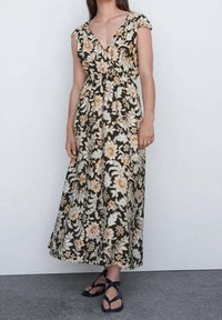 Maxikleid aus dunkelgrünem Stoff mit gelbem und weißem Blumenmuster, V-Ausschnitt, kurzen Ärmeln und tailliertem Schnitt; kombiniert mit schwarzen Sandalen.