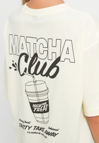 Kremfarget oversized T-skjorte med et grafisk design som viser ordene "MATCHA Club" og en drikkeillustrasjon i svart blekk.