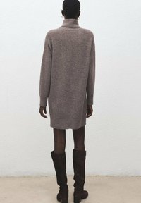 Pletené, oversized svetr šaty ve světle hnědé barvě s vysokým límcem. Nabízí dlouhé rukávy a boční rozparky na lemu, doplněné nadkolenkovými botami.