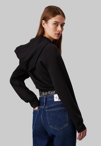 Čierna skrátená mikina s chrbtovým švom a kapucňou, doplnená vysokými modrými džínsami s viditeľným pásom Calvin Klein.