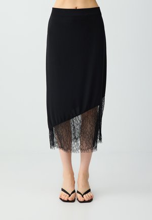 Jupe midi noire avec ourlet asymétrique et panneau en dentelle transparente, portée avec des sandales noires à brides et bout carré.
