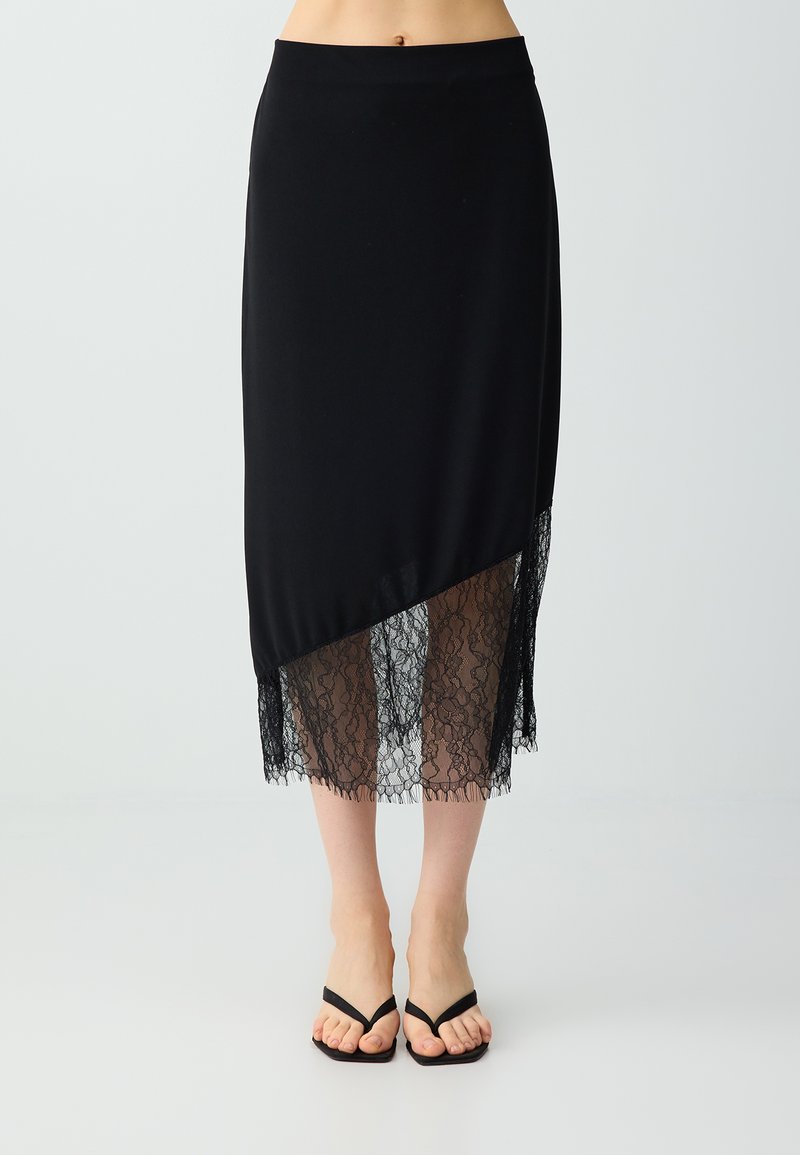 Jupe midi noire avec ourlet asymétrique et panneau en dentelle transparente, portée avec des sandales noires à brides et bout carré.