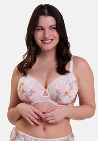 Reggiseno in pizzo floreale di un delicato pesco con rifiniture in pizzo bianco. Presenta motivi floreali e un dettaglio a fiocco al centro. Le spalline regolabili migliorano la vestibilità.