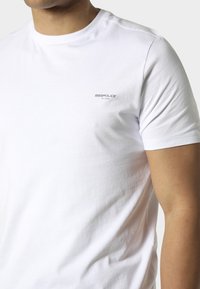 Uomo che indossa una t-shirt bianca a maniche corte con il logo "883 Police Milano" sul petto a sinistra, su sfondo semplice.