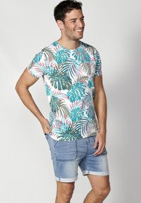 Camisa de manga corta para hombre con un estampado de hojas tropicales en verde, azul y rosa sobre un fondo blanco. Combinada con pantalones cortos de mezclilla.