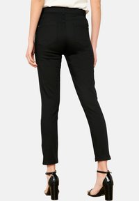 Juodi liekni džinsai su aukštu juosmeniu, subtiliomis užpakalinėmis kišenėmis ir lygia tekstūra, derinami su juodais aukštakulniais šlepetėmis.