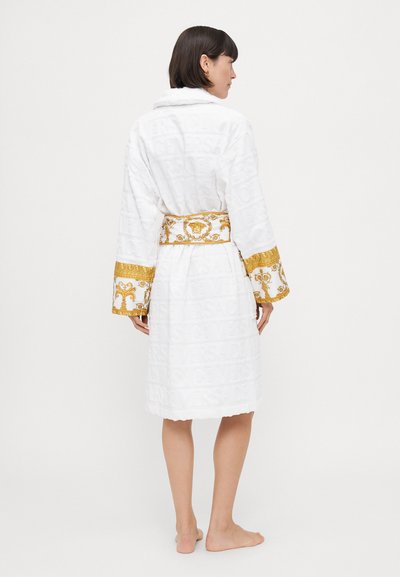 Rochie albă dintr-un material texturat, cu un logo embosat, curea aurie cu model și manșete decorative.
