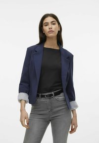 Mörkblå blazer med en strukturerad design och uppvikta randiga manschetter, bärs över en svart topp och kombineras med grå jeans. Minimalistisk stil.