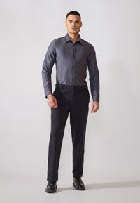 Camma da uomo grigia aderente con bottoni e una leggera lucentezza, abbinata a pantaloni eleganti neri e scarpe nere. Design semplice e slanciato con maniche lunghe.