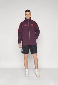 Giacca sportiva bordeaux con accenti rossi, chiusura a zip e logo. Indossata con pantaloni corti neri. Scarpe bianche e arancioni completano l'outfit.