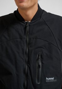 Mężczyzna w czarnej kurtce hummel na zamek błyskawiczny z pionową kieszenią na piersi i prążkowanym kołnierzem, widoczny od szyi do środka tułowia.