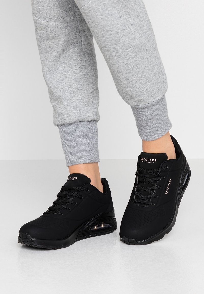 Black sports sneakers with textured fabric and air cushioning, featuring a lace-up design and silver "SKECHERS" branding on the tongue.