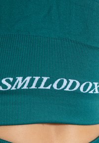 Tejido de canalé en color verde azulado con el texto "SMILODON" en blanco. Presenta un ajuste ceñido y una textura suave. El diseño enfatiza la comodidad y un aspecto moderno.