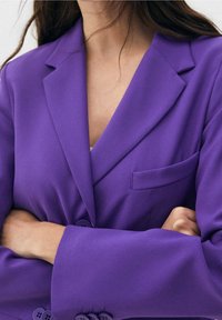 Blazer violet à la coupe structurée, avec revers crantés, une poche poitrine simple et de gros boutons sur les poignets. Texture de tissu lisse.
