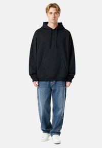 Felpa nera con cappuccio, tasca frontale e polsini a costine, abbinata a jeans larghi azzurri e sneaker bianche.