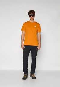 Oranž värvi lühikeste varrukatega t-särk väikese valge logoga, millele on lisatud mustad püksid ja mustad matkakotid, millel on tekstuuriga pind.