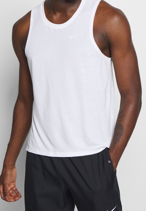 Sportlicher Mann trägt ein weißes, ärmelloses Nike-Tanktop und schwarze Nike-Shorts, eine Hand in der Tasche, vor neutralem Hintergrund.