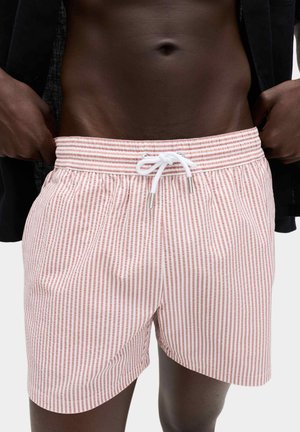 Męski tors w różowo-białych kąpielówkach w pionowe paski z białym sznurkiem, ręce regulujące czarną, otwartą koszulę.