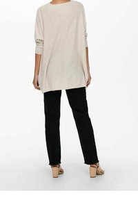 Personne portant un haut beige à manches longues ample, un pantalon noir à jambes droites avec des ourlets effilochés, et des sandales beiges à talons blocs, debout de dos.