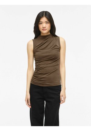 Jeune femme aux longs cheveux foncés portant un haut marron sans manches froncé et un pantalon noir, debout devant un fond blanc uni.