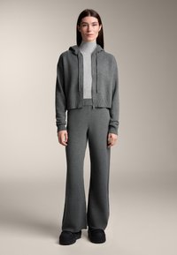 Hanorac gri, scurt, cu șnururi de ajustare și manșete striate, asortat cu pantaloni evazați, confecționați dintr-o țesătură moale tricotată. Pantofi negri cu platformă.