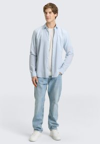 Jonge man die staat met één hand in zijn zak, gekleed in een lichtblauw open overhemd over een wit T-shirt, losse lichtblauwe jeans en witte sneakers.