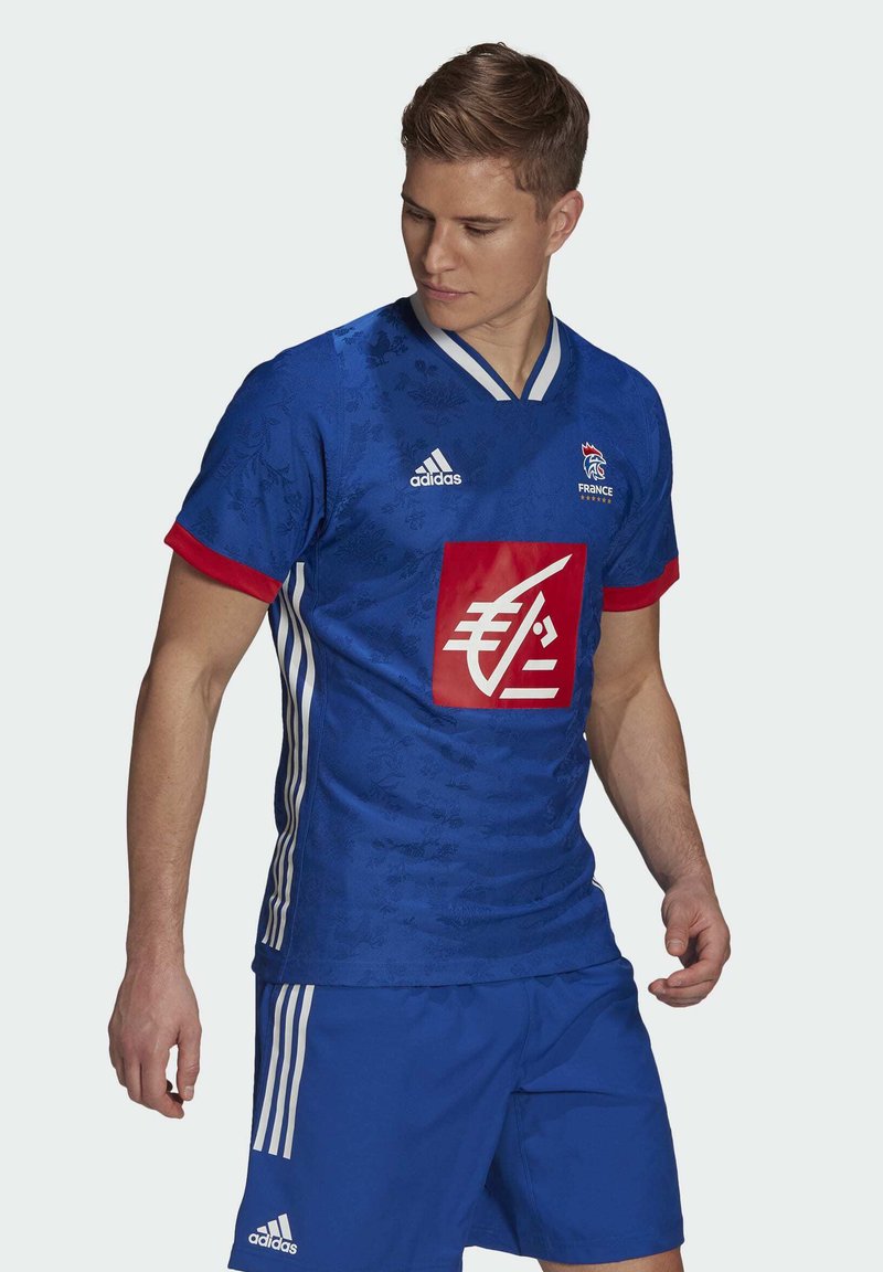 Blauw voetbalshirt met een bloemenpatroon, rode accenten en een centraal logo. Heeft witte strepen op de mouwen en shorts.