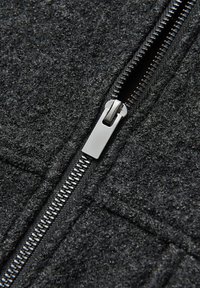 Tessuto grigio di lana con una cerniera argentata lungo la cucitura, che mostra una superficie strutturata con fibre visibili e dettagli di cucitura.