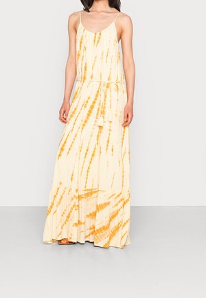 Kvinde iført en ærmeløs maxi-kjole med tie-dye i creme og orange med tynde stropper og et bælte i taljen med kvaster, stående mod en ensfarvet baggrund.