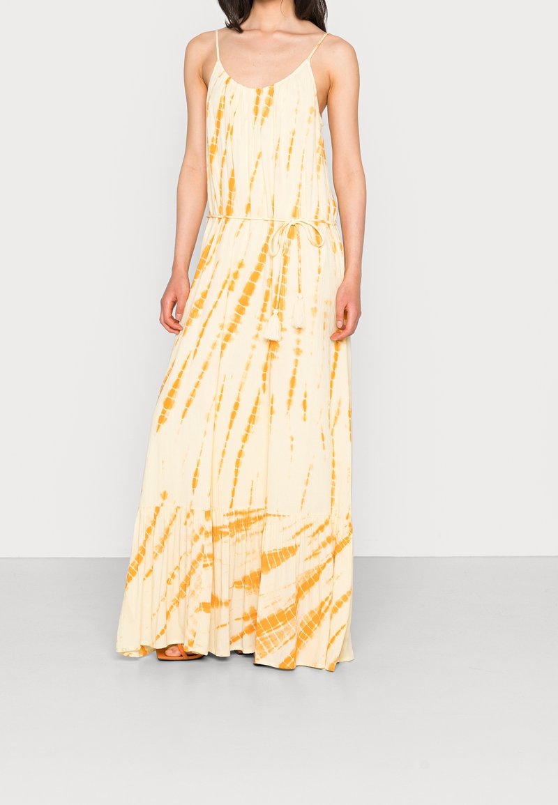 Femme portant une robe longue sans manches tie-dye crème et orange avec de fines bretelles et une ceinture à la taille avec des pompons, debout devant un fond uni.