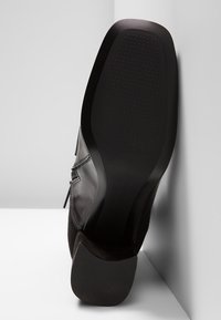 Chaussure habillée en cuir noir montrée de côté, avec un léger motif texturé et un petit talon sur un fond gris en coin.