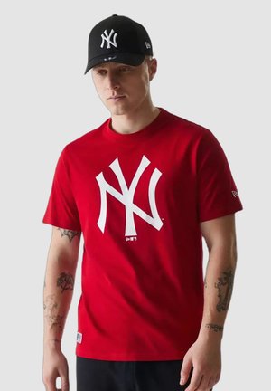 Hombre con camiseta roja con un gran logo blanco y gorra de béisbol negra, de pie frente a un fondo liso gris claro.