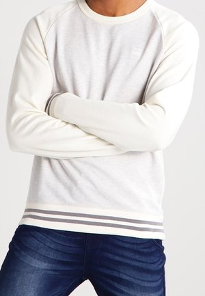 Sudadera gris claro con mangas raglán crema y dobladillo a rayas; presenta una textura suave y un detalle de logo en el pecho. Combinada con jeans de mezclilla.