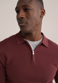 Vino crvena pletena polo majica s ovratnikom na patentni zatvarač, s teksturiranim tkanjem, preko sive majice. Krupni plan gornjeg dijela tijela i lica.