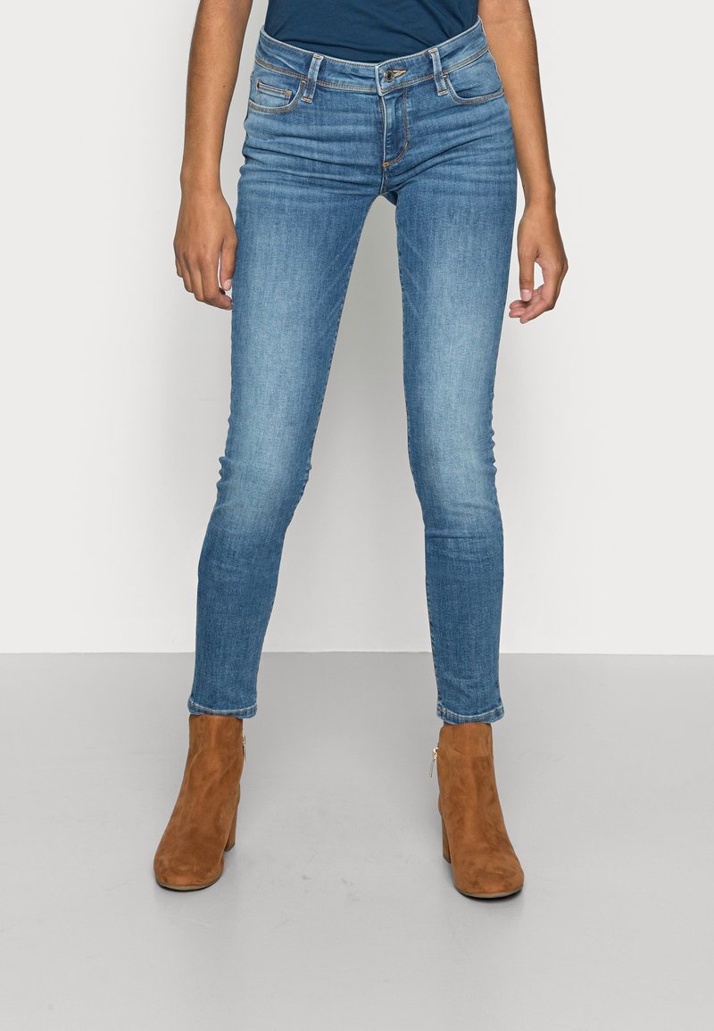 Štíhlé modré džínsy vyrobené z denimové tkaniny s mírným vyblednutím, zásluhou pětisloupcového designu a poutka na opasek, doplněné hnědými semišovými kotníkovými botami.