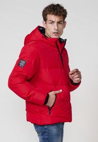 Chaqueta acolchada roja con capucha, cierre de cremallera y bolsillos laterales. Presenta detalles en negro y un acabado texturizado. El modelo muestra un perfil lateral.