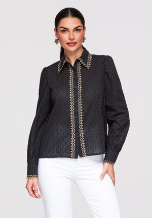 Frau trägt eine schwarze langärmelige Bluse mit goldener Einfassung am Kragen, vorne und an den Manschetten, kombiniert mit weißen Hosen vor einem schlichten hellen Hintergrund.