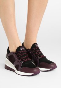 Fötter i maroon och svarta sneakers med vita sulor, med snören och nätpaneler på sidorna mot en enkel ljus bakgrund.
