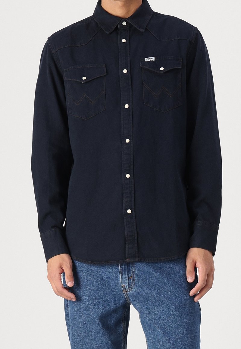 Homme portant une chemise bleu marine foncé à manches longues avec deux poches sur la poitrine et un jean bleu clair sur un fond uni.