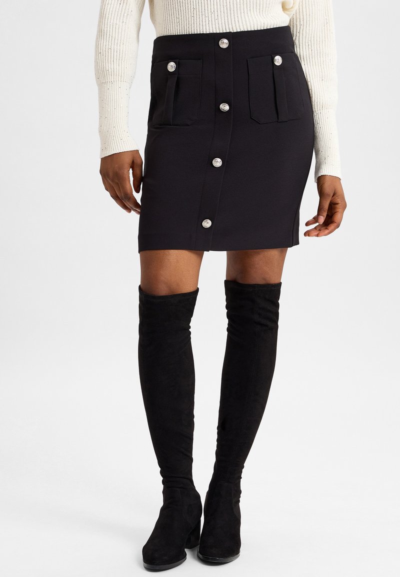 Schwarzer, taillierter Rock mit vorderer Knopfleiste und zwei seitlichen Taschen. Kombiniert mit schwarzen Wildleder-Stiefeln bis zum Oberschenkel. Texturiertes cremefarbenes Sweatshirt darüber.