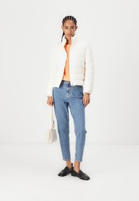 Hvit quiltert pufferjakke, oransje genser, blå jeans, svarte mokkasiner, og en liten beige håndveske. En enkel hvit bakgrunn.
