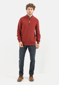 Roter Strickpullover mit einem Viertelreißverschluss, strukturiertem Muster, gerippten Bündchen, kombiniert mit dunklen Jeans und braunen Schnürstiefeln.