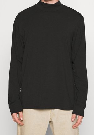 Homme portant un col roulé noir à manches longues et un pantalon beige, debout devant un fond clair uni.