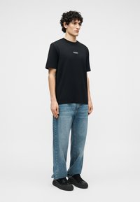 Hombre con cabello rizado que lleva una camiseta negra con logo, jeans azules holgados y zapatillas negras, de pie contra un fondo claro y liso.