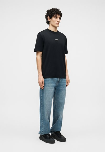 Homme aux cheveux bouclés portant un T-shirt noir avec logo, un jean bleu ample, et des baskets noires, debout devant un fond clair uni.